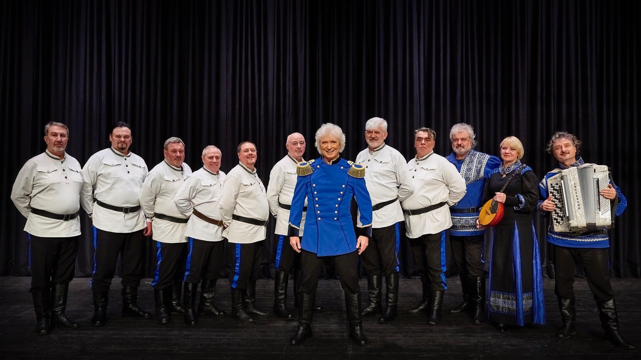 Peter Orloff in blauer Uniform und elf Mitgliedern des Kosaken Chores in weißen Tuniken vor dunklem Vorhang