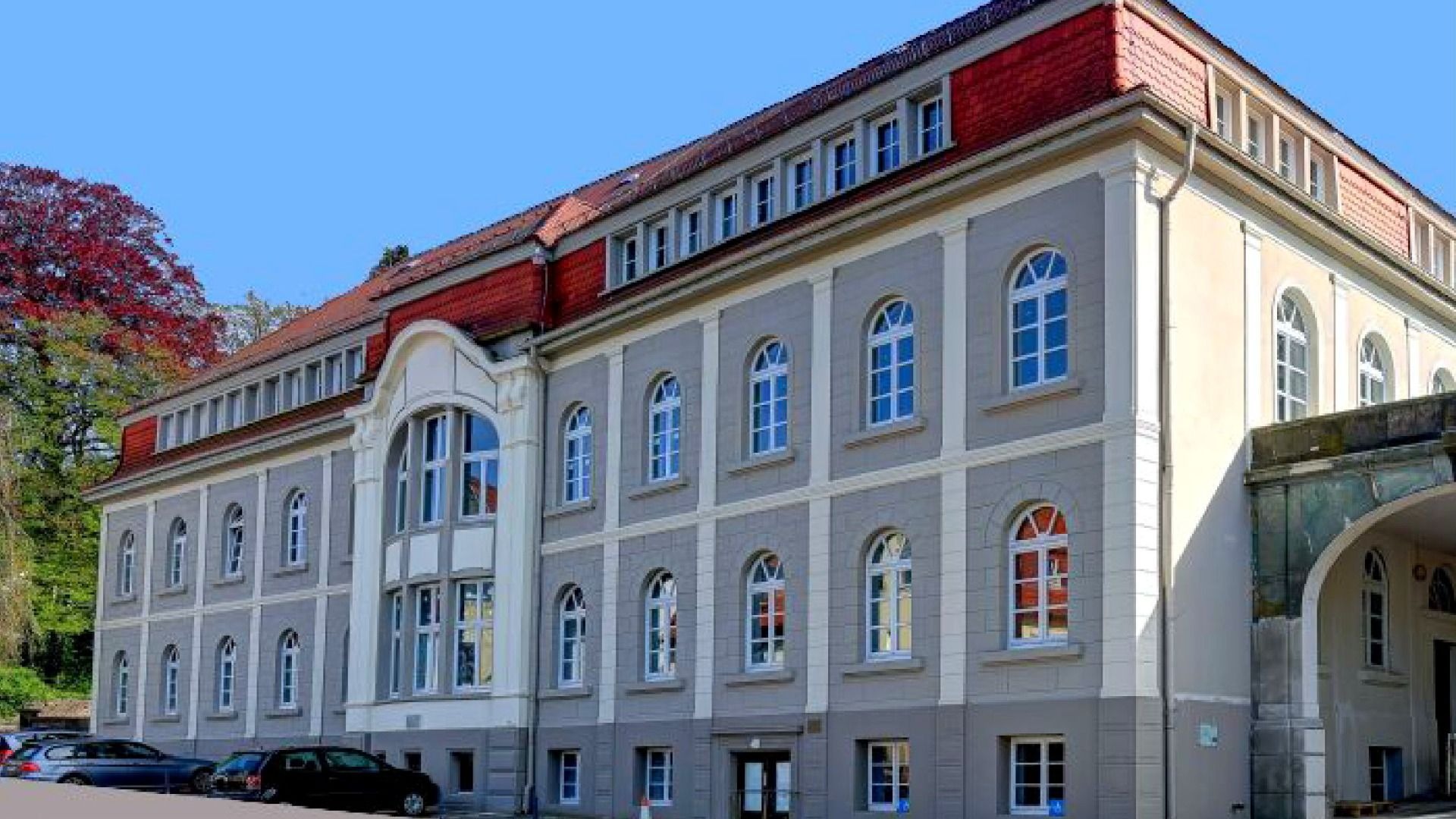 Großes, graues und beiges Gebäude mit rotem Dach, vielen Bogenfenstern und geparkten Autos