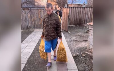 Junger Mann trägt zwei Netze voller Kartoffeln auf einem gepflasterten Weg, dahinter geht eine weitere Person