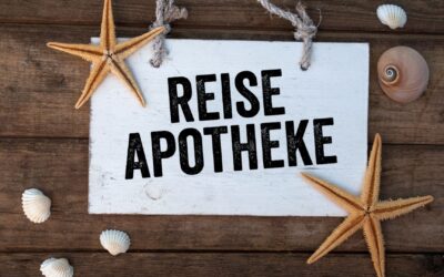 Weißes Holzschild mit der Aufschrift "REISE APOTHEKE" auf braunem Holzuntergrund, umgeben von Seesternen und Muscheln