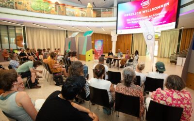 Publikum lauscht Podiumsdiskussion des Festivals „Denken Fühlen Handeln“ vor großem „Herzlich Willkommen“-Bildschirm