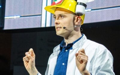 Mann in Laborkittel und auffälligem gelben Helm mit Mikrofon spricht und gestikuliert auf Bühne