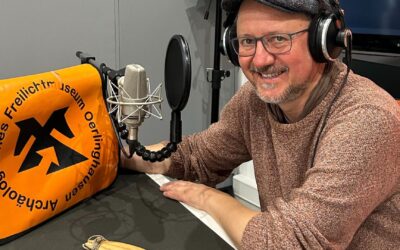 Lächelnder Mann mit Kopfhörern und Mikrofon in einem Aufnahmestudio neben einer Tasche des Museums Oerlinghausen