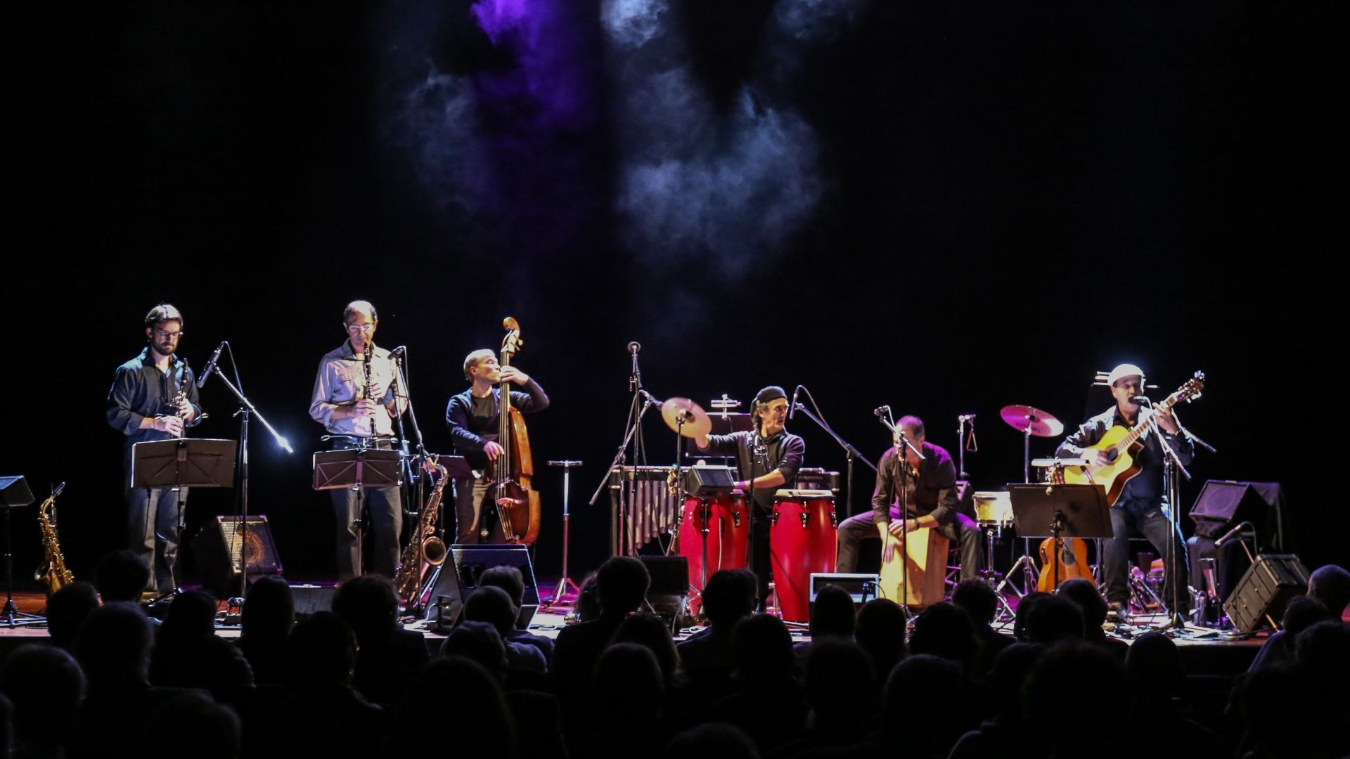 Sechs Musiker spielen auf einer dunklen Bühne Gitarre, Blasinstrumente, Kontrabass und Perkussion vor einem Publikum