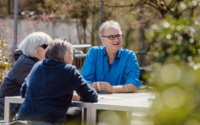 Drei Personen sitzen an einem Tisch im Freien, ein Mann in blauem Hemd lacht und unterhält sich mit zwei anderen