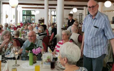 Eine Gruppe Senioren sitzt an Tischen in einem Restaurant, ein Mann mit Brille und Hemd steht im Gang
