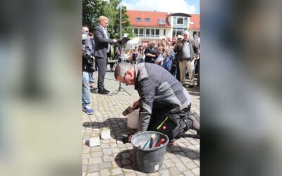 Mann verlegt Stolpersteine auf gepflastertem Platz, während ein Redner spricht und Menschen zuschauen
