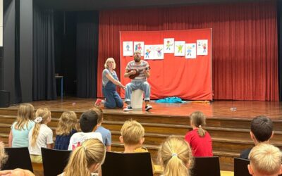 Mann spielt Ukulele und Frau kniet auf Bühne vor Kindern, Hintergrund rote Vorhänge mit vielen Kinderzeichnungen