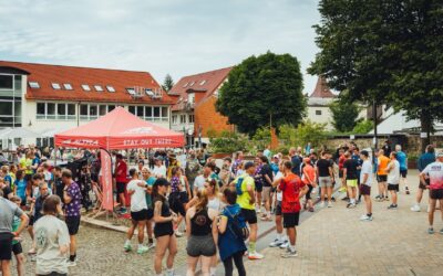 Zahlreiche Läufer und Besucher stehen auf einem Stadtplatz, umgeben von Häusern mit roten Dächern und einem roten