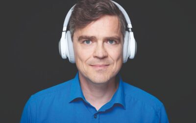 Henning Schmidtke lächelt, trägt ein blaues Hemd und weiße Kopfhörer auf dunklem Hintergrund