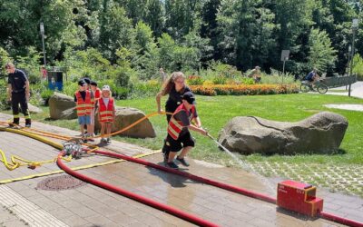 Kinderfeuerwehrübung: Ein Kind spritzt mit einem Wasserschlauch auf ein rotes Feuerwehrmodell, andere Kinder schauen zu