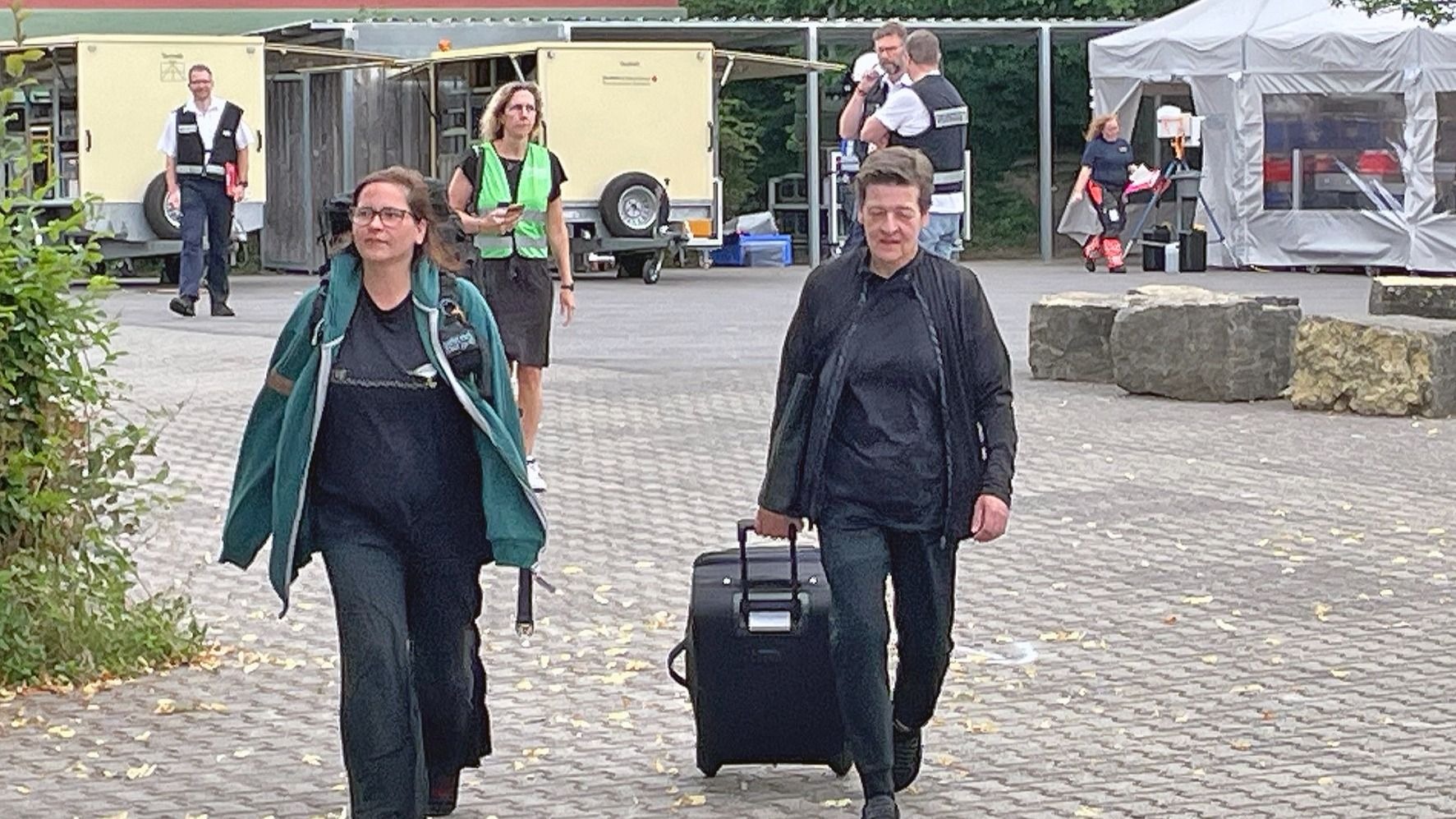 Zwei Frauen gehen auf gepflastertem Boden, eine zieht Koffer, mehrere Menschen in Warnwesten stehen zwischen Zelten und