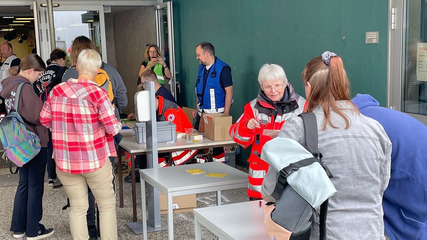 Mehrere Menschen, darunter Einsatzkräfte in roten Jacken und blauen Westen, stehen an Tischen bei einer Registrierung
