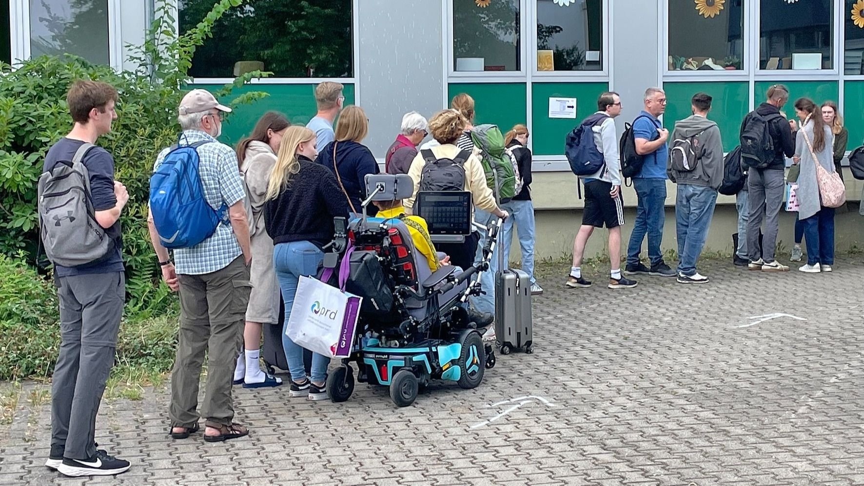 Eine Menschengruppe mit Rucksäcken und einer Person im Elektrorollstuhl wartet vor einem grünen Gebäude auf