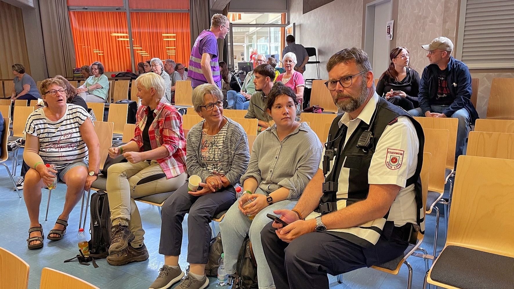 Ein Feuerwehrmann mit Funkgerät und weitere Personen sitzen in Reihen auf Holzstühlen in einem Saal
