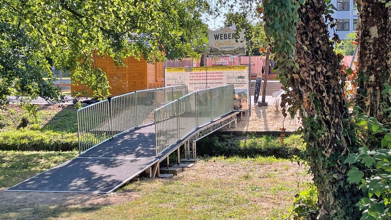 Eine temporäre Fußgängerbrücke führt zu einem Parkbereich mit Schildern für Webers Garten und Hortus Vitalis