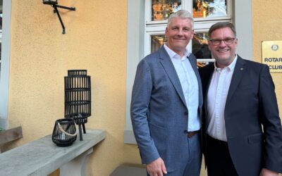 Zwei lächelnde Männer in dunklen Anzügen, einer mit Brille, stehen vor gelber Hauswand und Rotary Club Schild
