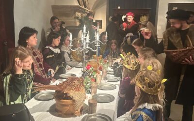 Kinder und Erwachsene in historischen Kostümen sitzen oder stehen an einer festlich gedeckten Tafel mit einem Fasan