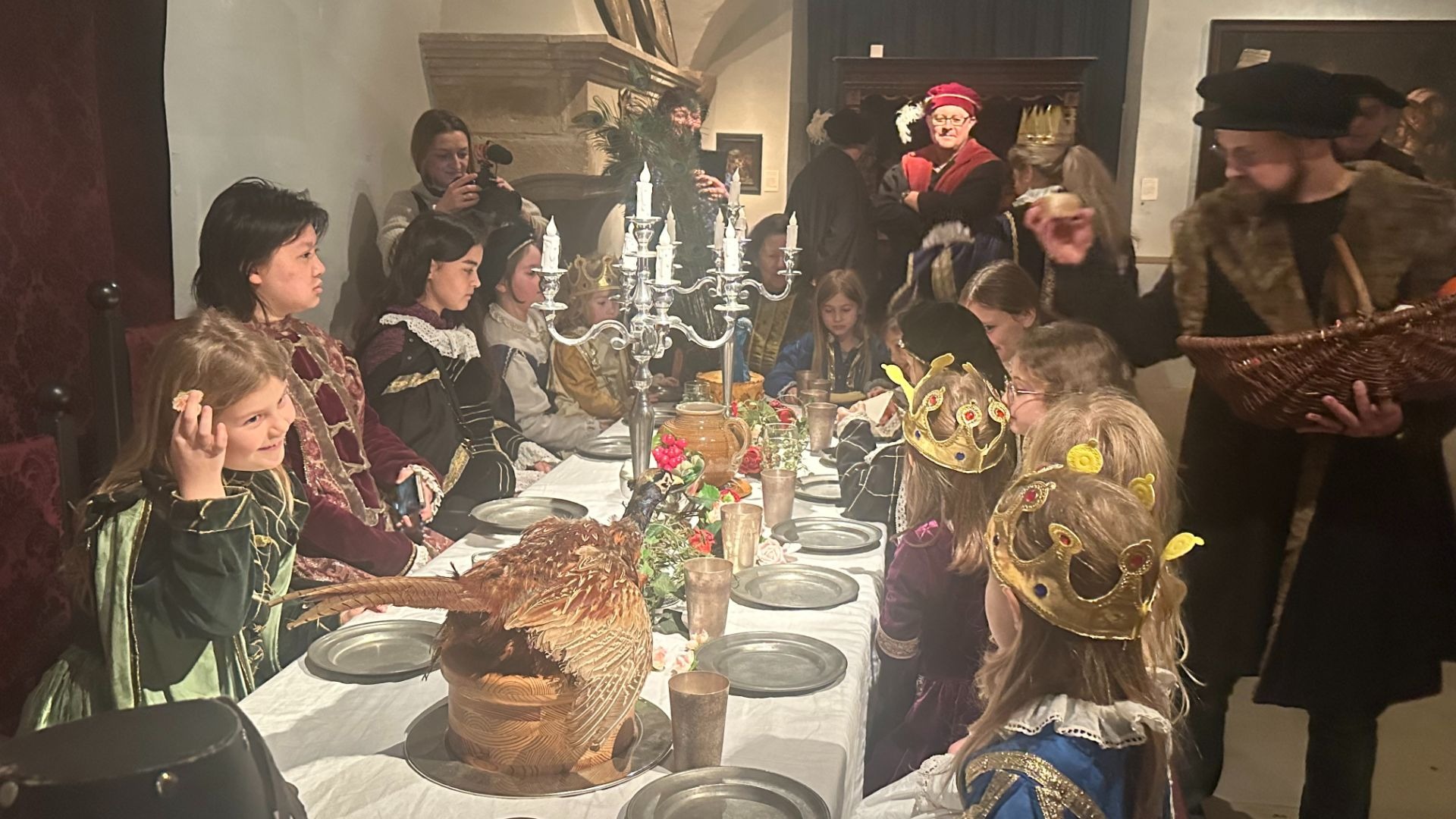 Kinder und Erwachsene in historischen Kostümen sitzen oder stehen an einer festlich gedeckten Tafel mit einem Fasan