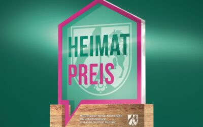 Hausförmiges 3D-Logo mit der Aufschrift HEIMAT PREIS und dem Wappen von Nordrhein-Westfalen auf einem Holzsockel