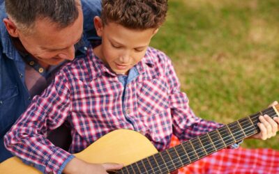 Junge spielt Gitarre, während ein Mann daneben auf einer karierten Decke im Gras sitzt