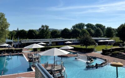 Sommerliches Freibad mit Sport- und Erlebnisbecken, weißen Sonnenschirmen, Umkleiden und grünen Bäumen an einem klaren