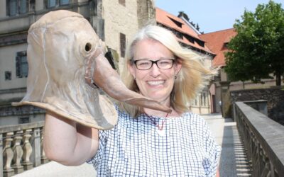Lächelnde Frau mit Brille hält große beigefarbene Pestmaske mit Schnabel vor historischer Steinfassade und