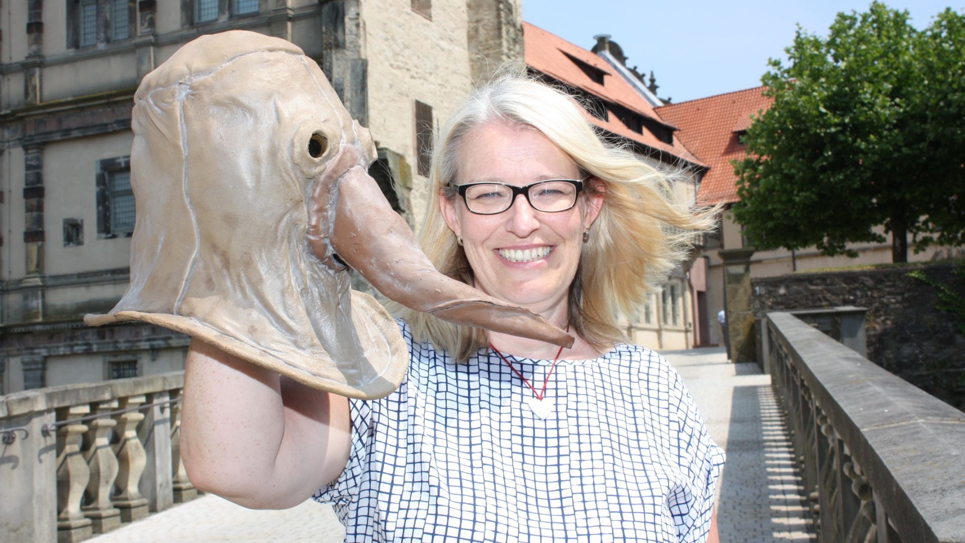 Lächelnde Frau mit Brille hält große beigefarbene Pestmaske mit Schnabel vor historischer Steinfassade und