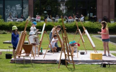 Fünf Kinder und eine erwachsene Frau malen an Staffeleien im Park vor einem Gebäude, weitere Personen im Hintergrund