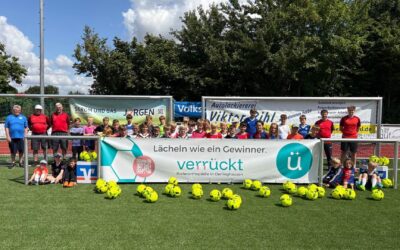 Kinder und Betreuer eines Fußballcamps posieren auf einem Fußballplatz mit vielen gelben Bällen und Sponsorbannern