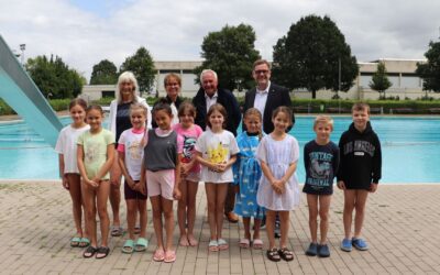 Zehn Kinder und sechs Erwachsene stehen am Beckenrand eines Freibads vor einer blauen Wasserrutsche