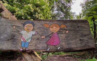 Holzschild mit aufgemalten, lächelnden Kindern, einem Jungen mit Brille und einem Mädchen mit Zöpfen, im Wald