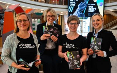 Vier lächelnde Frauen präsentieren in einem modernen Gebäude Programmhefte der GENIALE 2026 "Wie geht's uns?