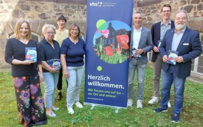Sieben lächelnde Personen stehen vor einem vhs-Banner und einer Steinwand, halten Broschüren "Wissen als Kompass für