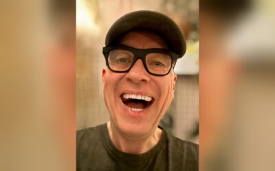 HG Butzko lacht herzlich mit offenem Mund, trägt dunkle Mütze und schwarze Brille