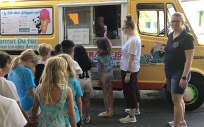 Personen, darunter viele Kinder, stehen vor einem gelben Eiswagen, ein Kind erhält eine Eistüte