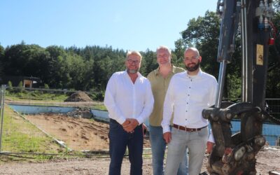 Drei Männer lächeln auf der Baustelle des Oerlinghauser Freibads mit abgerissenem Becken und Wald
