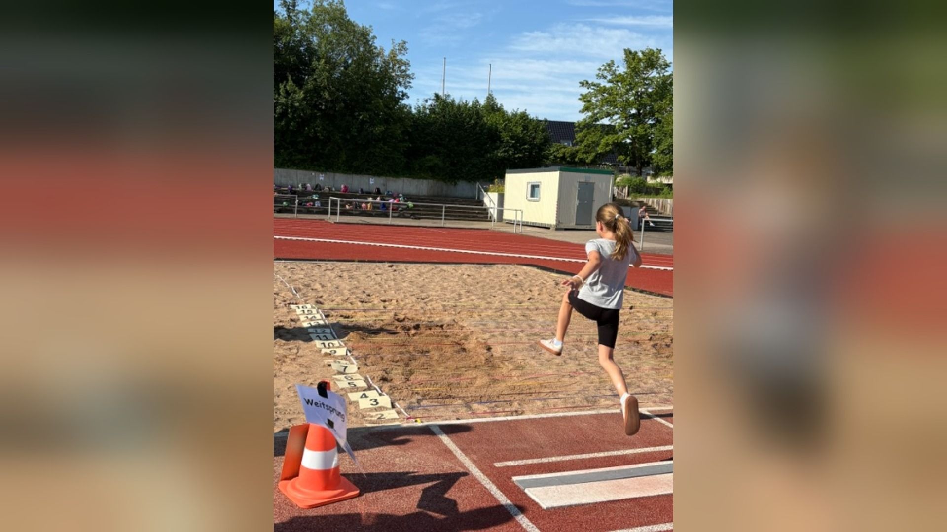 Mädchen springt gerade in eine Weitsprunggrube auf einer Sportanlage mit roter Laufbahn und Tribüne im Hintergrund