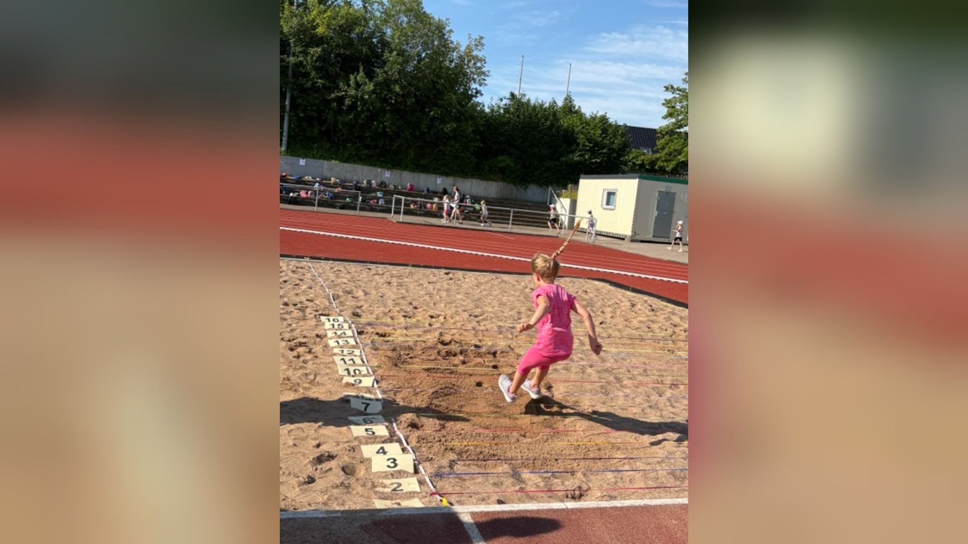 Mädchen in rosa Kleidung springt in eine Sandgrube beim Weitsprung auf einem Sportplatz, dahinter Tribüne und Laufbahn