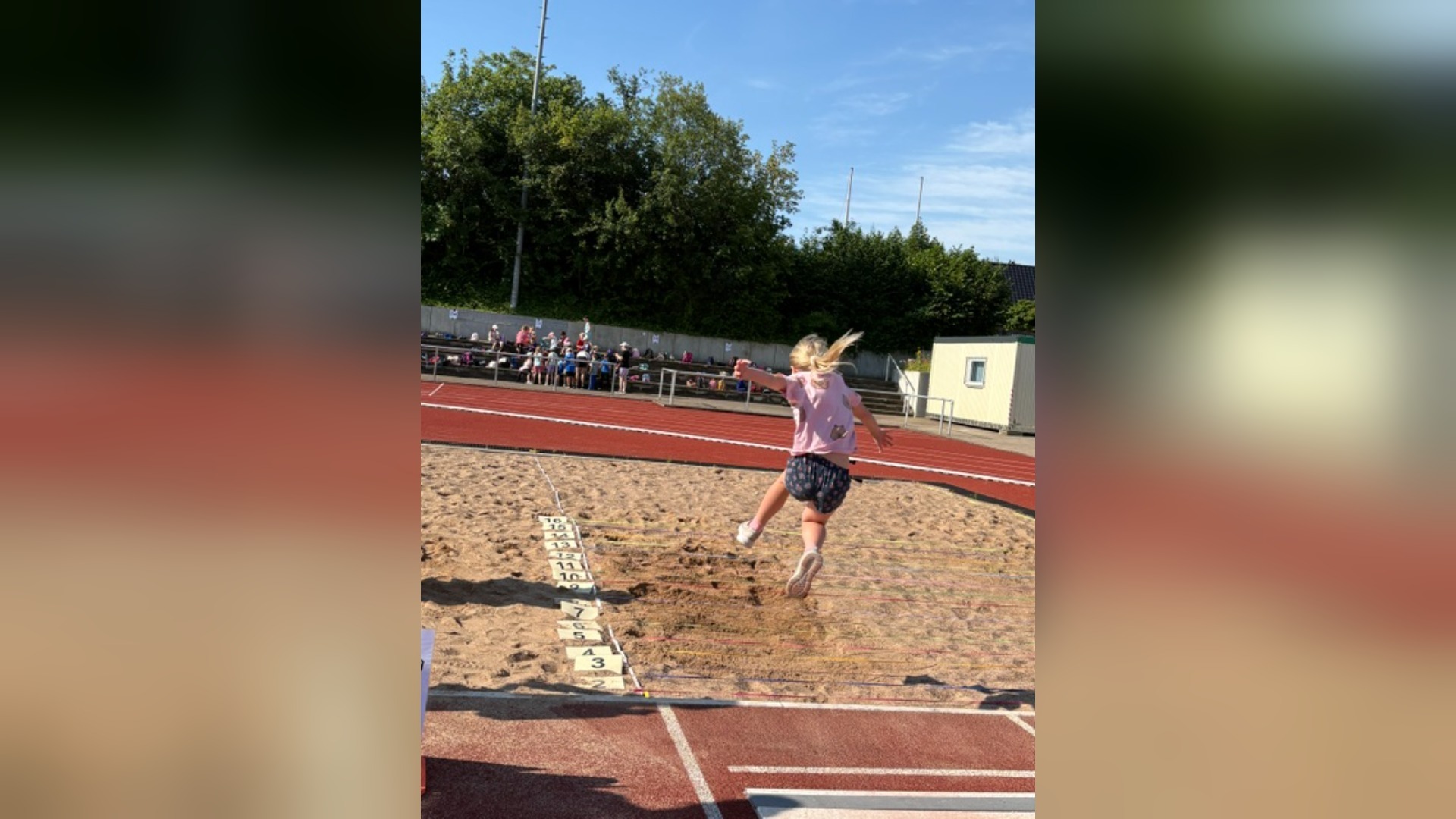 Junges blondes Mädchen beim Weitsprung in die Sandgrube, im Hintergrund sitzen Zuschauer auf der Tribüne