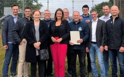 Zehn lächelnde Personen, zwei Frauen und acht Männer, stehen vor einem Zaun, einer hält einen Förderbescheid