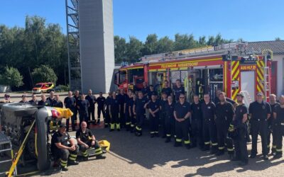 32 Feuerwehrleute aus Augustdorf posieren vor ihrem Löschfahrzeug und einem auf die Seite gelegten Auto