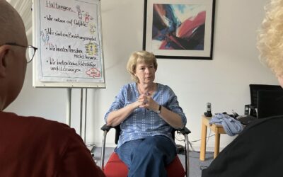 Drei Personen in einem Raum, eine Frau spricht, während zwei andere zuhören, hinter ihr ein Flipchart mit Überschrift