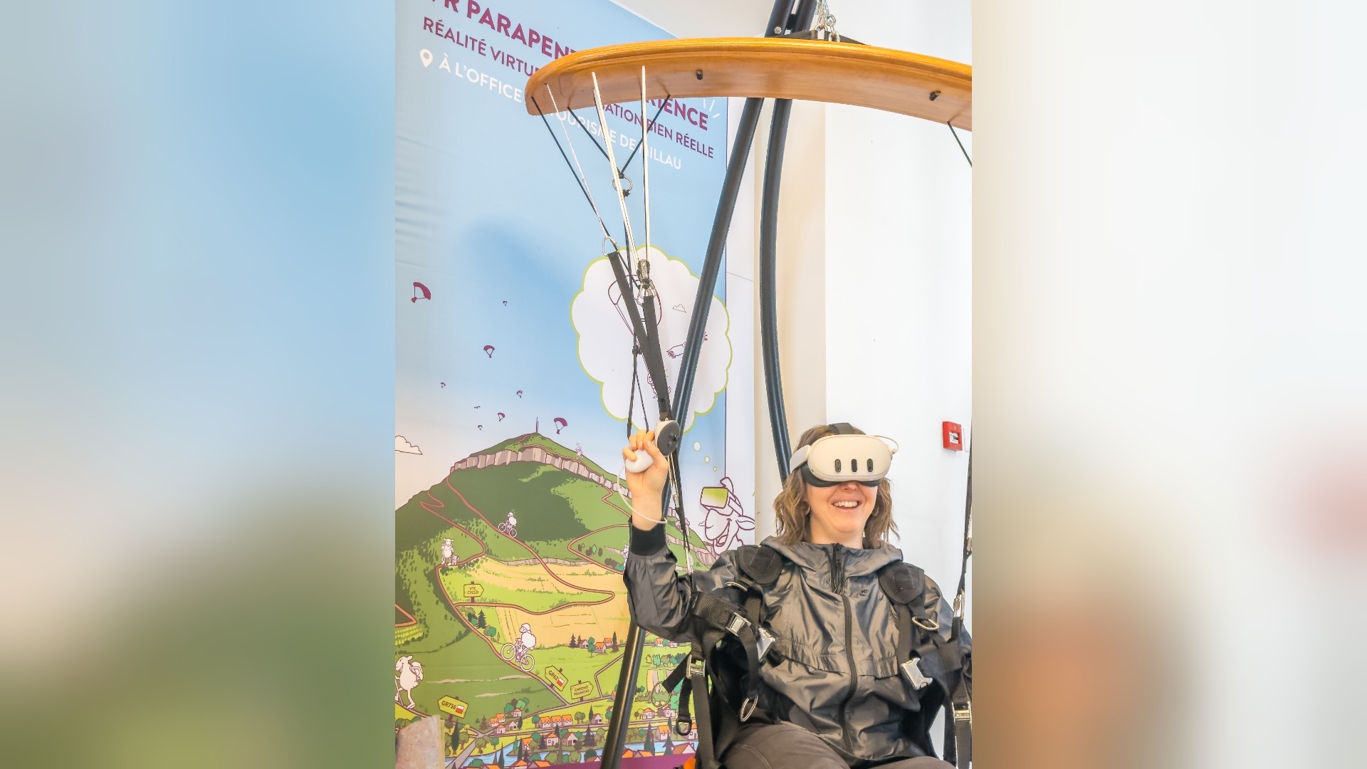 Lächelnde Frau mit VR-Brille sitzt in Paragliding-Simulator vor einer illustrierten Berglandschaft mit Millau