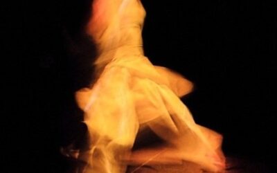 Eine in gelbem Licht dynamisch tanzende Person mit fließendem, hellem Kleid vor dunklem Hintergrund