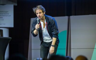 Ein junger Mann mit Bart und Schal lehnt sich sprechend mit Mikrofon auf einer Bühne nach vorne