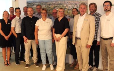 Elf lächelnde Männer und Frauen stehen in einem Raum vor einer großen Wandgrafik einer Siedlung