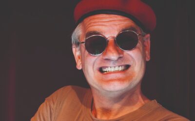 Mann mit rotem Barett und runder Sonnenbrille lächelt breit mit verschränkten Armen vor dunklem Hintergrund
