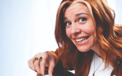 Laura Brümmer mit roten Haaren, brauner Cordjacke und Goldschmuck, die lächelnd direkt in die Kamera schaut