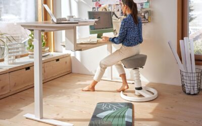 Frau arbeitet auf ergonomischem Hocker an einem Schreibtisch mit zwei Arbeitsplatten und Computern vor einem Fenster